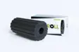 Sort foam roller med dybe langsgående riller hele vejen rundt, stillet foran en hvid Blackroll Groove-æske